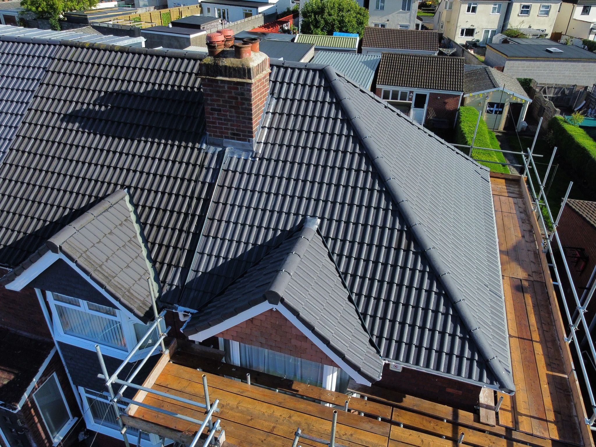 Aerial view of a house with new dark gray roof tiles and a red brick chimney. Scaffolding surrounds the building, indicating ongoing construction or renovation. Nearby rooftops and a green lawn are visible.