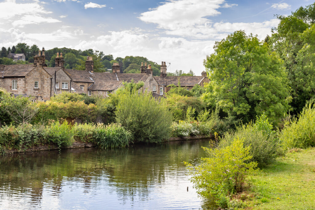 Stone cottages with chimneys line a lush, green riverbank. Bushes and trees border the calm water under a partly cloudy sky, creating a peaceful rural scene.