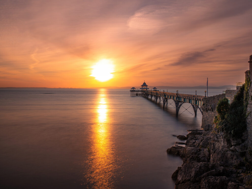 A long pier extends over calm water at sunset, with the orange sun low on the horizon and its reflection shimmering on the sea. Rocky shoreline and soft-colored clouds frame the tranquil scene.