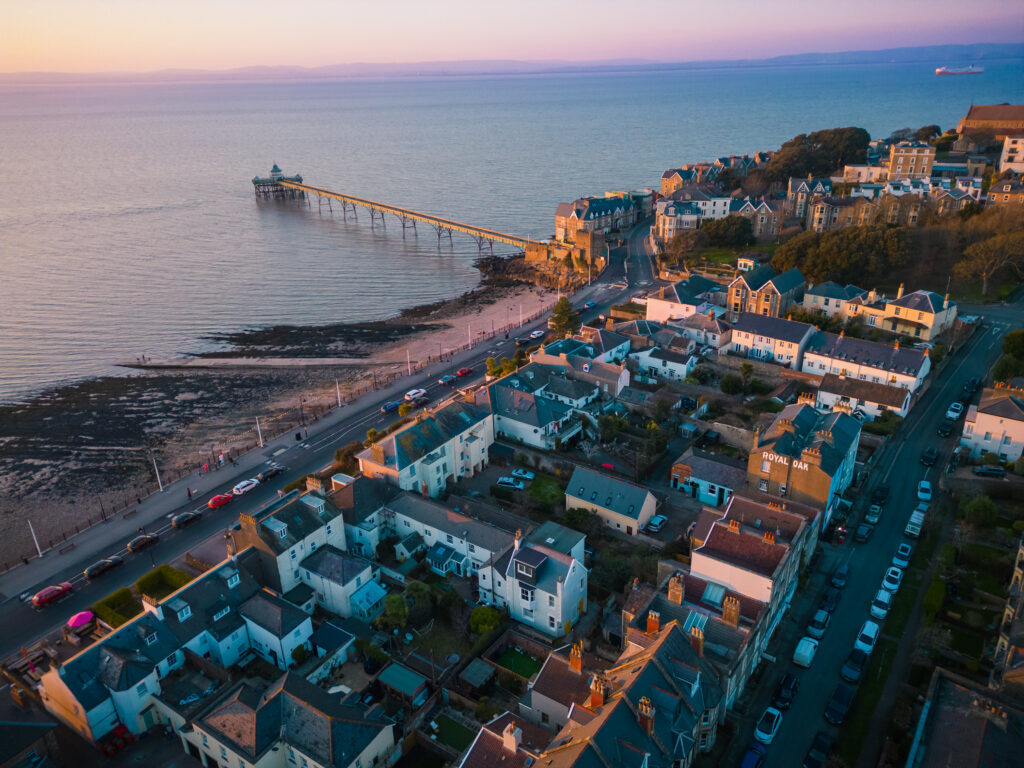 Aerial view of a coastal town at sunset with houses, a long pier stretching into the calm sea, and a shoreline with parked cars and people walking along the beach.