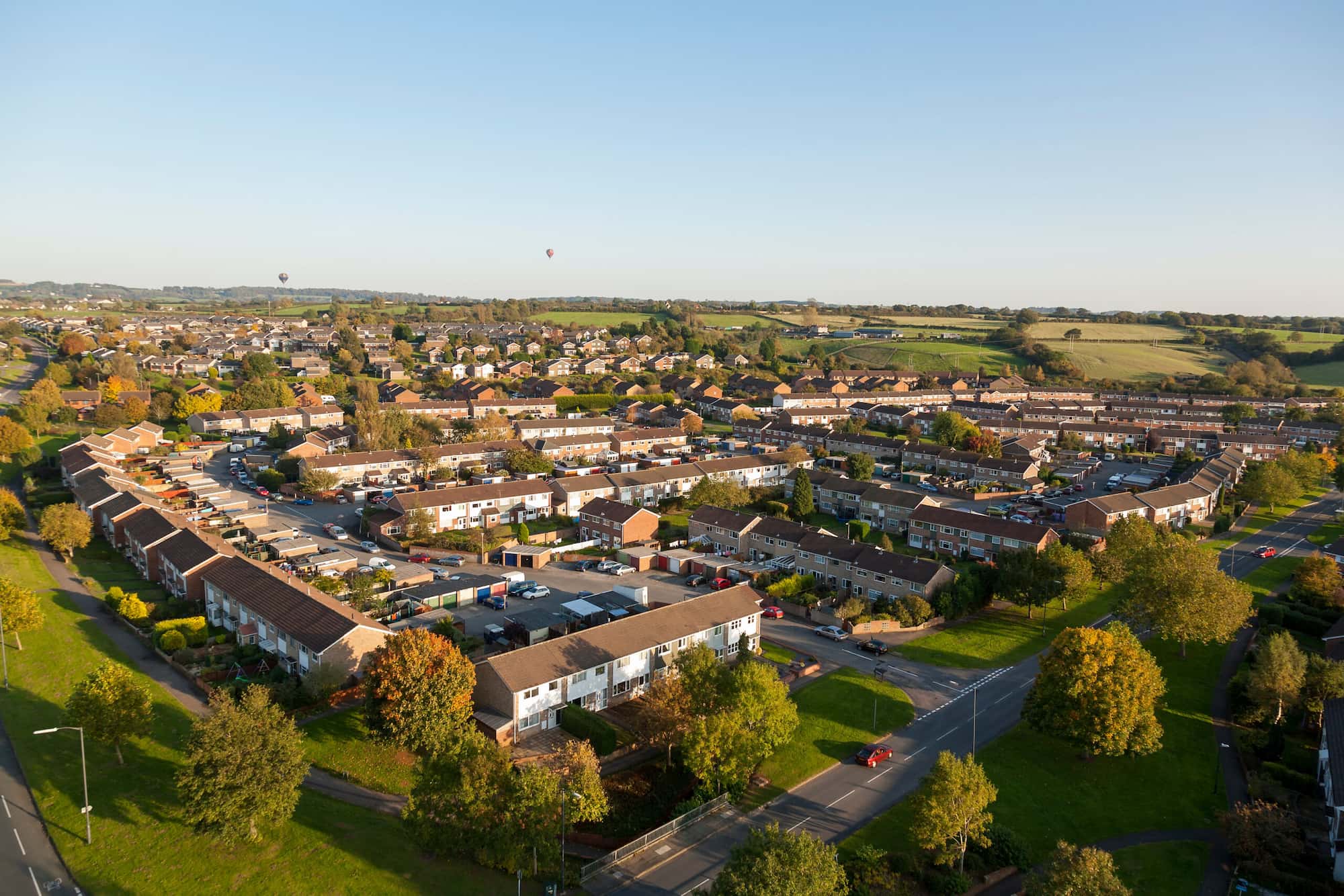 Aerial view of a suburban neighborhood with rows of houses, green trees, parked cars, and open fields in the background under a clear blue sky.